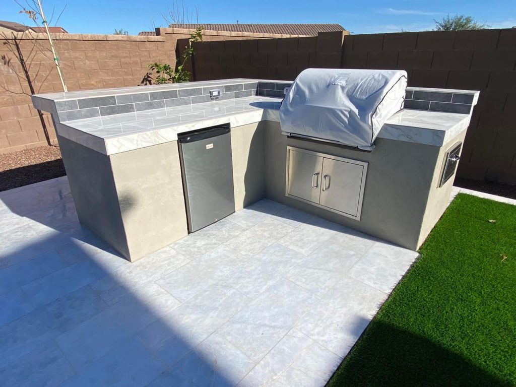An outdoor kitchen setup with a built-in barbecue grill covered with a protective cover. To the left of the grill, there's a countertop that likely serves as a preparation area and below it is what appears to be a compact refrigerator. On the right-hand side, there's a storage cabinet under the counter. The flooring is a combination of tiles and artificial turf, providing a clean and maintenance-friendly surface. The walls surrounding the area are high, offering privacy for the space.