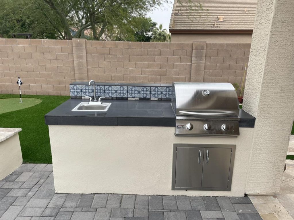 Outdoor kitchen with a built-in gas grill, sink, and storage on a patio with patterned tile flooring. A concrete block wall surrounds the area.