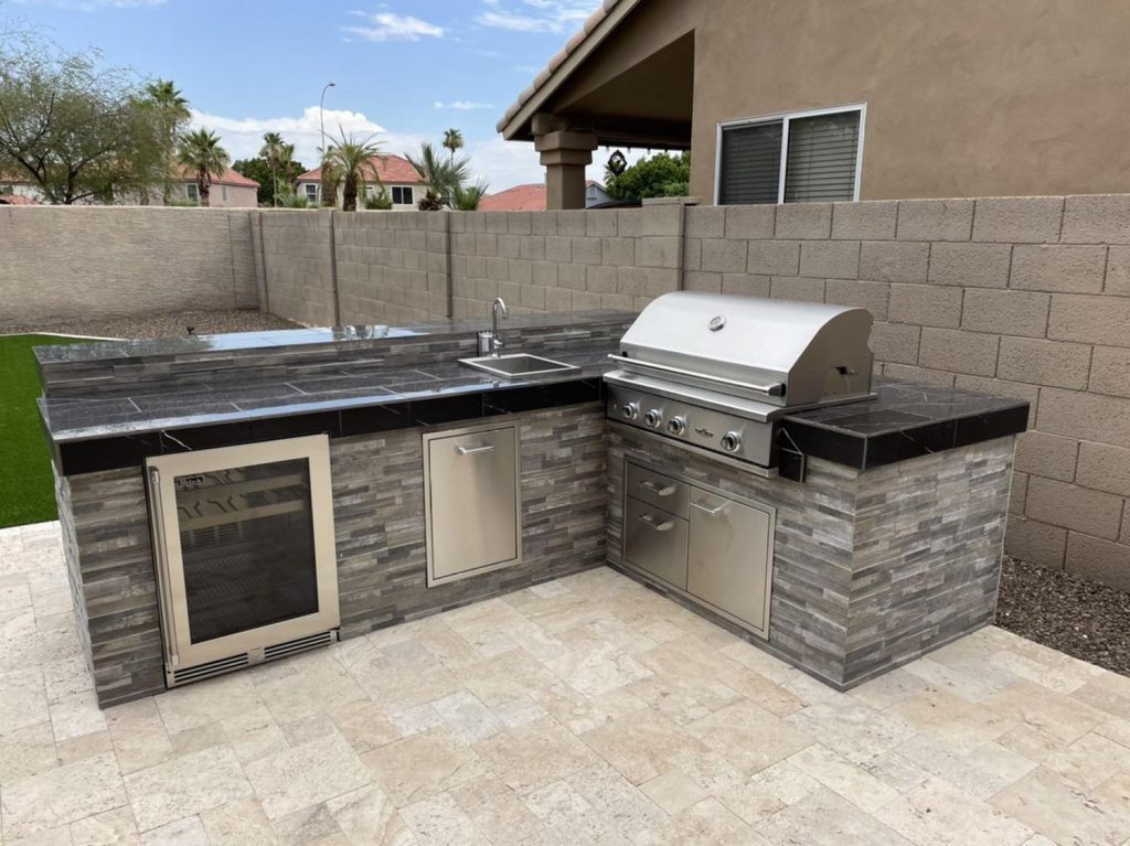 A sophisticated outdoor kitchen featuring a grill, refrigerator, and cabinetry set against a stone facade under a covered patio with polished stone flooring. A wall encloses the backyard.