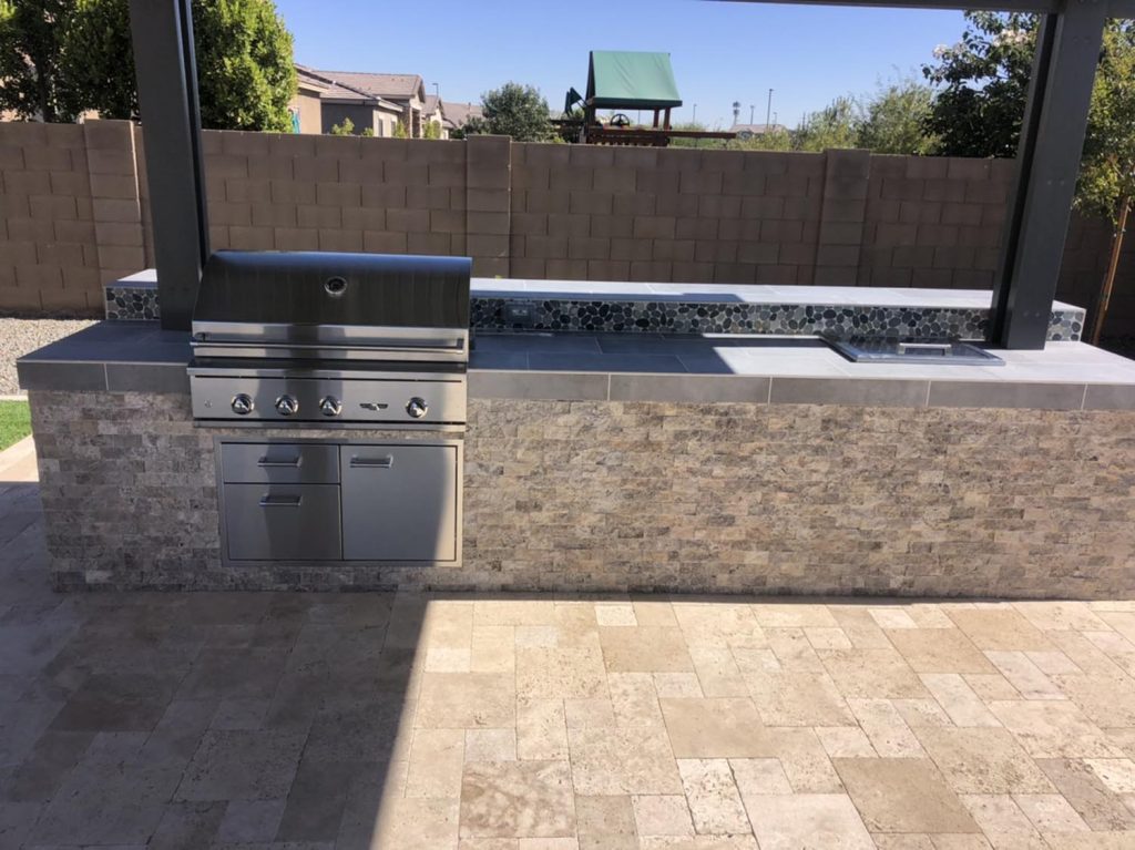 An outdoor grilling station featuring a stainless steel barbecue grill, storage drawers, and a sink, with a tile backsplash and a paved patio.