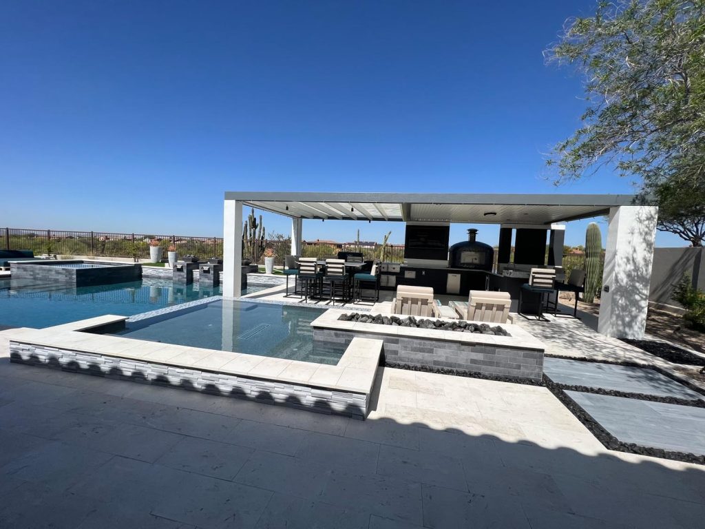 Luxurious outdoor living space featuring a covered patio with furniture and a built-in pizza oven, a reflective pool with stepping stones, surrounded by desert landscape and a clear blue sky.