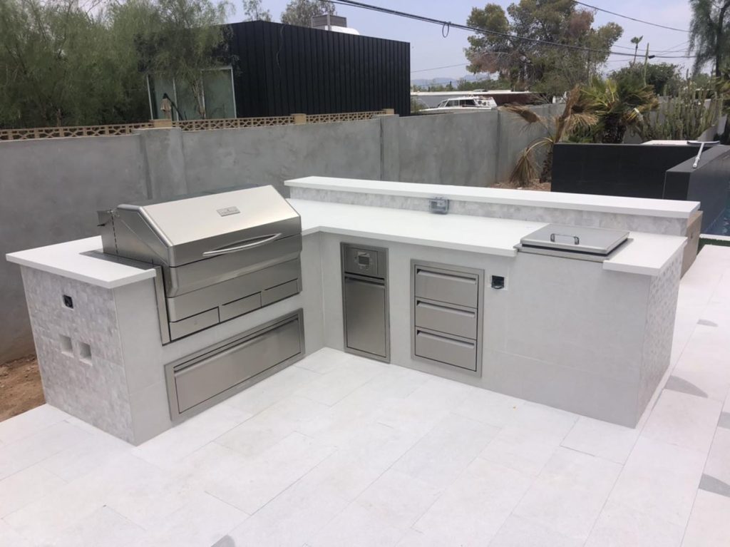 A modern outdoor kitchen setup with a stainless steel grill, multiple storage compartments, and a white countertop, against a concrete wall and a tiled floor.