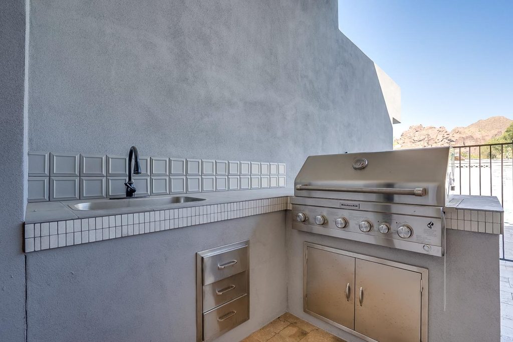 Close-up of an outdoor kitchen area featuring a stainless steel grill with knobs and a storage cabinet below. To the left is a sink with a black faucet, set against a wall with white square tiles and a white tiled countertop.