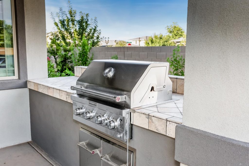 An outdoor kitchen with a built-in barbecue grill on a tiled counter, adjacent to the exterior wall of a home.
