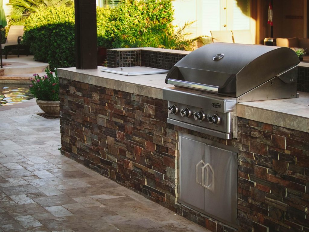 A stainless steel barbecue grill on a stone counter of an outdoor kitchen, with a landscaped yard and pool area visible in the background.