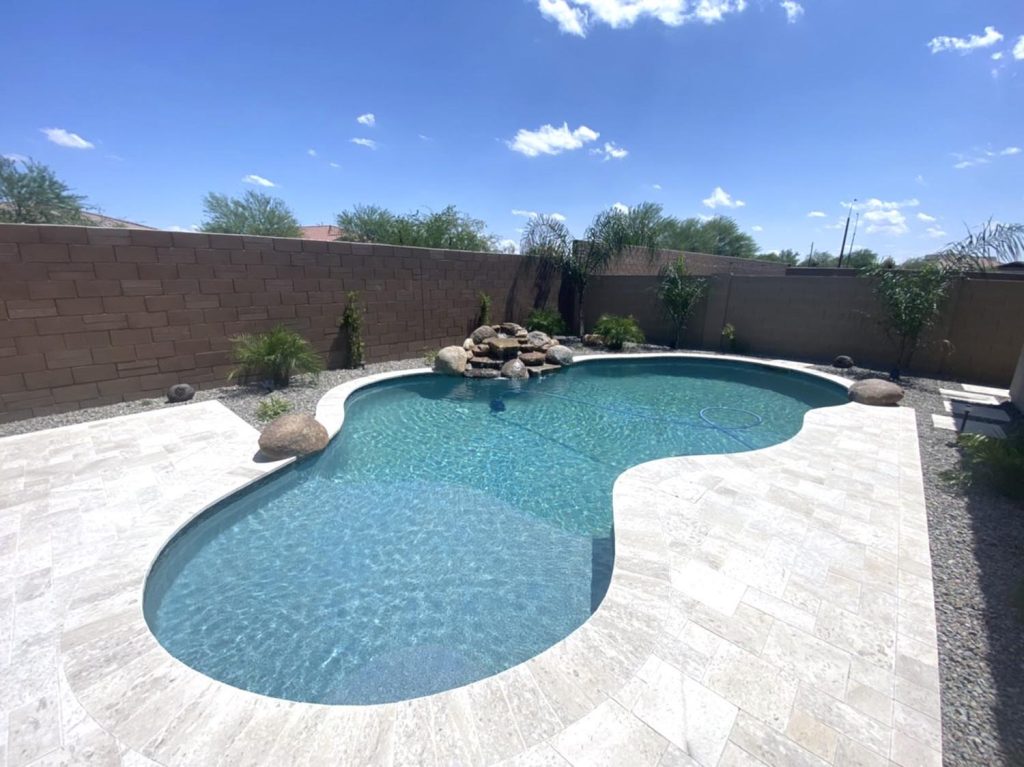 A curvilinear residential swimming pool with clear blue water, bordered by a pale stone deck. The pool is adorned with a cluster of rocks on one side from which water appears to flow. Several plants, both tall and short, dot the perimeter. A tall brown brick wall encloses the backyard, and the sky above is bright with scattered clouds.