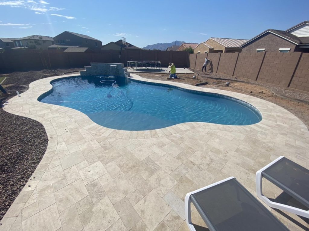 A sunlit residential swimming pool with a curvilinear shape, surrounded by a light-colored stone patio. Houses with brown roofs are visible in the background. Workers are present at the site, finalizing construction. A distant mountain range is visible against a clear blue sky.