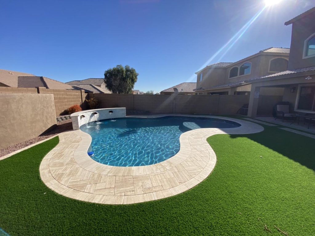 Set against a clear blue sky, the pool exhibits a neat design with a blend of curved and straight edges. Surrounding the pool is a bright artificial turf, contrasting nicely with the pool's deep blue. The residential setting is evident from the houses in the background, and the high walls provide privacy for those enjoying a dip. A simple water feature arches into the pool, adding a touch of elegance.