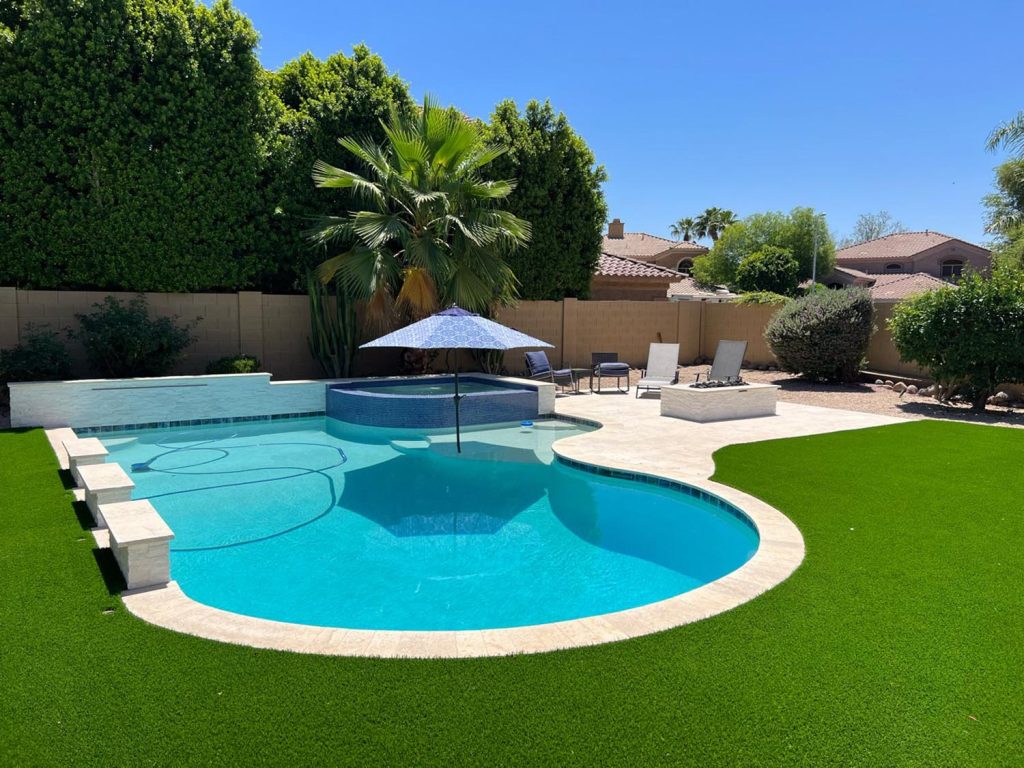 This pool setting offers a luxurious vibe. A striking feature is the raised, transparent section of the pool, giving an illusion of floating water. Sun loungers and outdoor furniture are set up for relaxation, and an umbrella provides shade. The pool is bordered by vibrant artificial turf and thick, dense shrubs. The surrounding houses indicate a upscale Arizona suburban setting, while the well-maintained yard suggests an upscale residence.