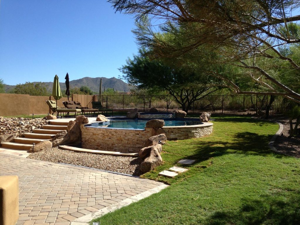 Daylight scene of a desert-themed backyard with a swimming pool encased in a stone structure, sun loungers, natural boulders, scattered gravel, grassy areas, and a fence with a mountainous view beyond.