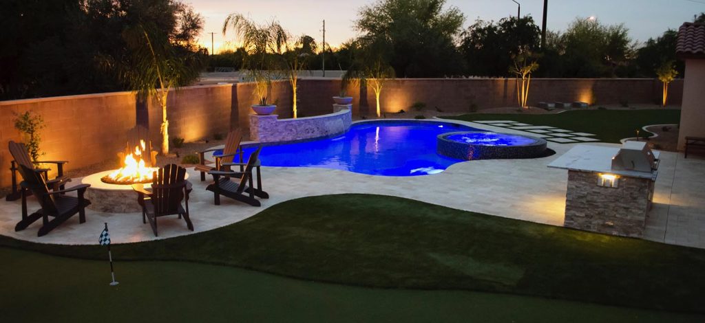Illuminated backyard at dusk with a blue swimming pool, fire pit surrounded by wooden chairs, adjacent stone-built jacuzzi, mini-golf setup on green turf, and an outdoor grill station with stone countertop.