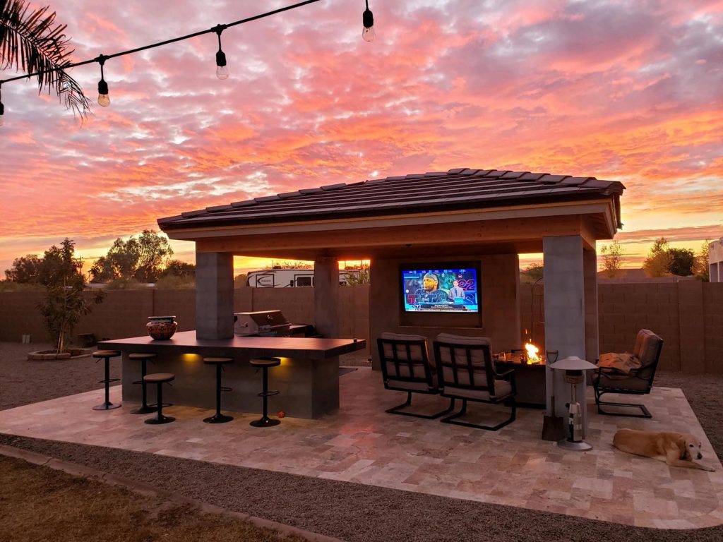 Outdoor residential area at sunset with a vibrant sky, featuring an outdoor kitchen with a TV, a fire pit, bar stools, and a resting dog.
