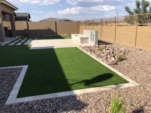 A residential backyard with synthetic turf sections bordered by white concrete and natural stones, with a pebble path leading to a brown wall with a built-in shelf and two structures resembling chimney outlets.
