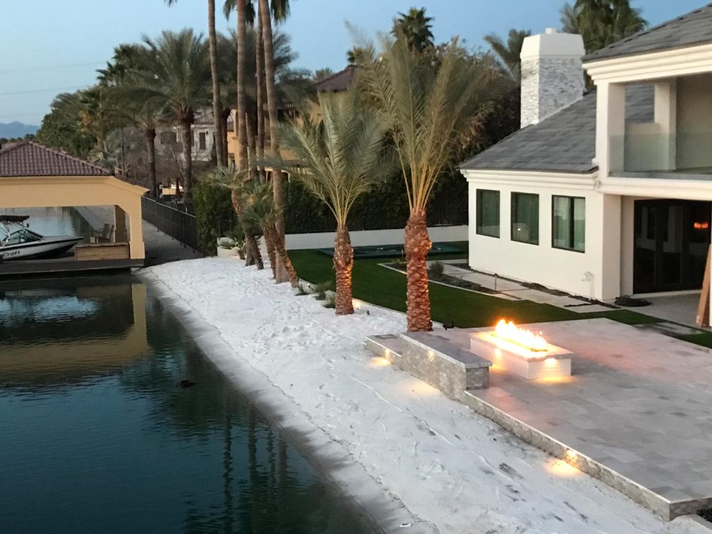 A residential outdoor area during dusk with palm trees, a lit fire pit on the shore, and a boat under a roofed structure on the water.