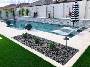 A modern backyard swimming pool with submerged loungers, artificial grass, decorative stones, and a striped umbrella, set against a privacy wall with planters.