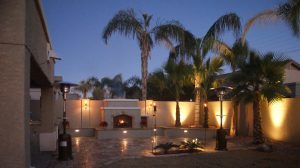 An evening view of a residential patio with a fireplace, palm trees, and outdoor lighting, with the sky transitioning from dusk to night.