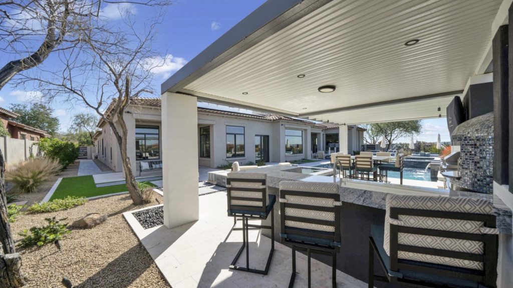 An outdoor covered patio with a ceiling fan and lights, featuring a bar area with stools by a poolside, artificial turf, and leafless trees in the background under a clear blue sky.