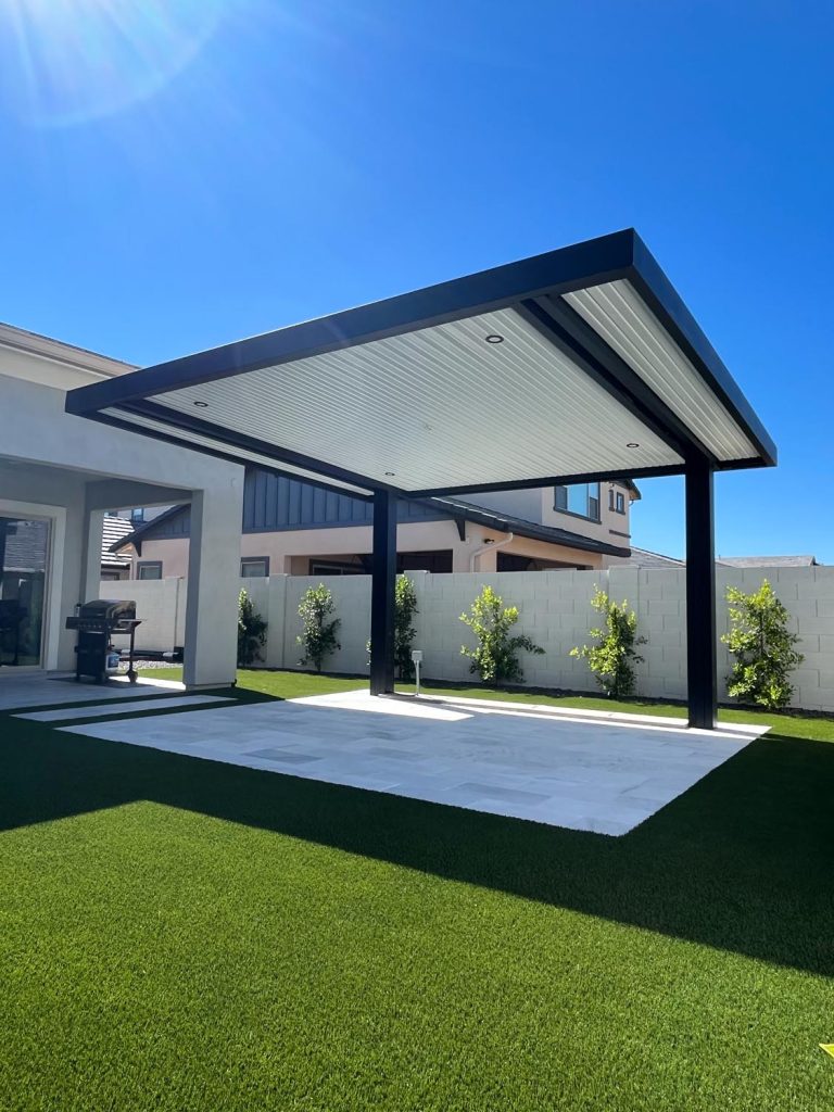 A modern backyard patio with a large, angular pergola providing shade over a neatly manicured artificial turf. The sky is clear and blue, indicating a sunny day. A barbecue grill is visible in the background, suggesting a space designed for outdoor leisure activities.