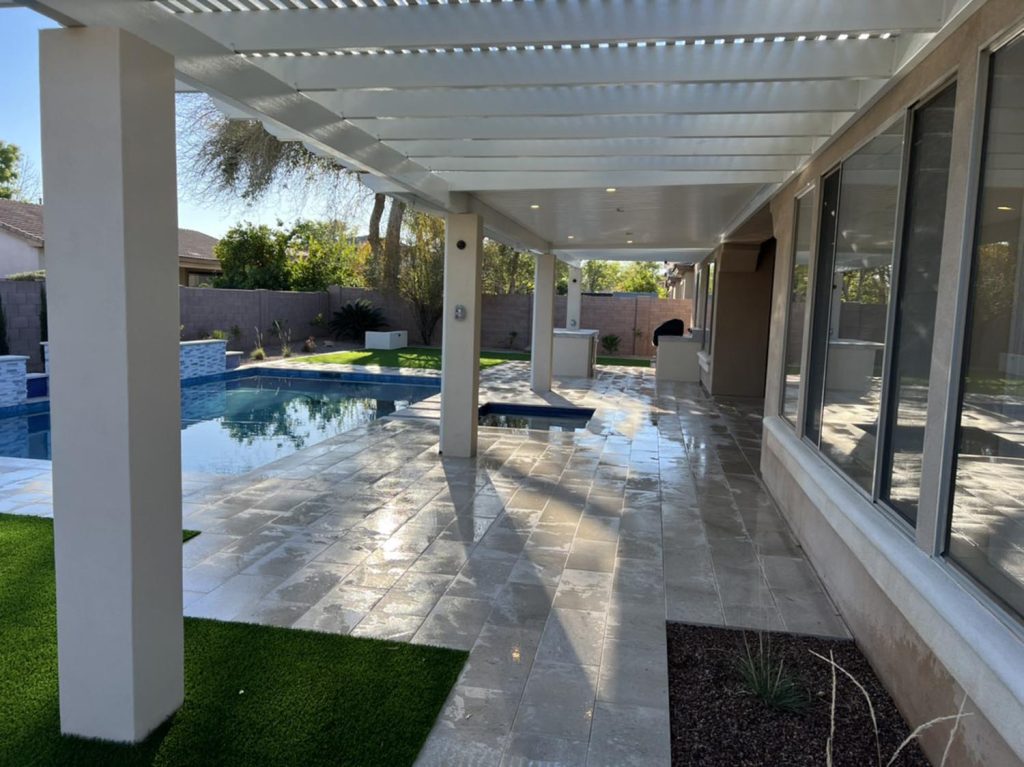 A sheltered outdoor patio area with a glossy tiled floor that reflects the sky above. The patio is adjacent to a pool with clear, calm water. Columns support the overhanging structure, which is equipped with louvered roof panels. The space appears to be a cool, shaded area for relaxation near the pool, with the boundary wall of the property in the distance.