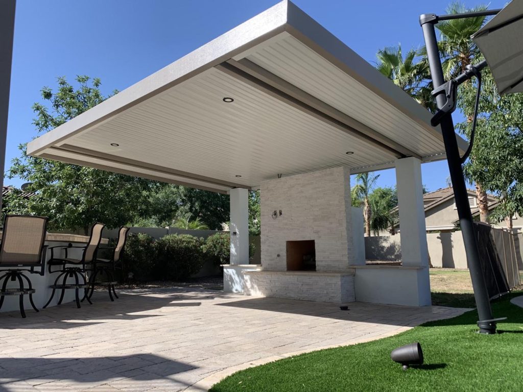 A simple covered outdoor patio area with a brick fireplace, paved flooring, outdoor furniture under a pergola with recessed lighting, surrounded by artificial grass and residential fencing.