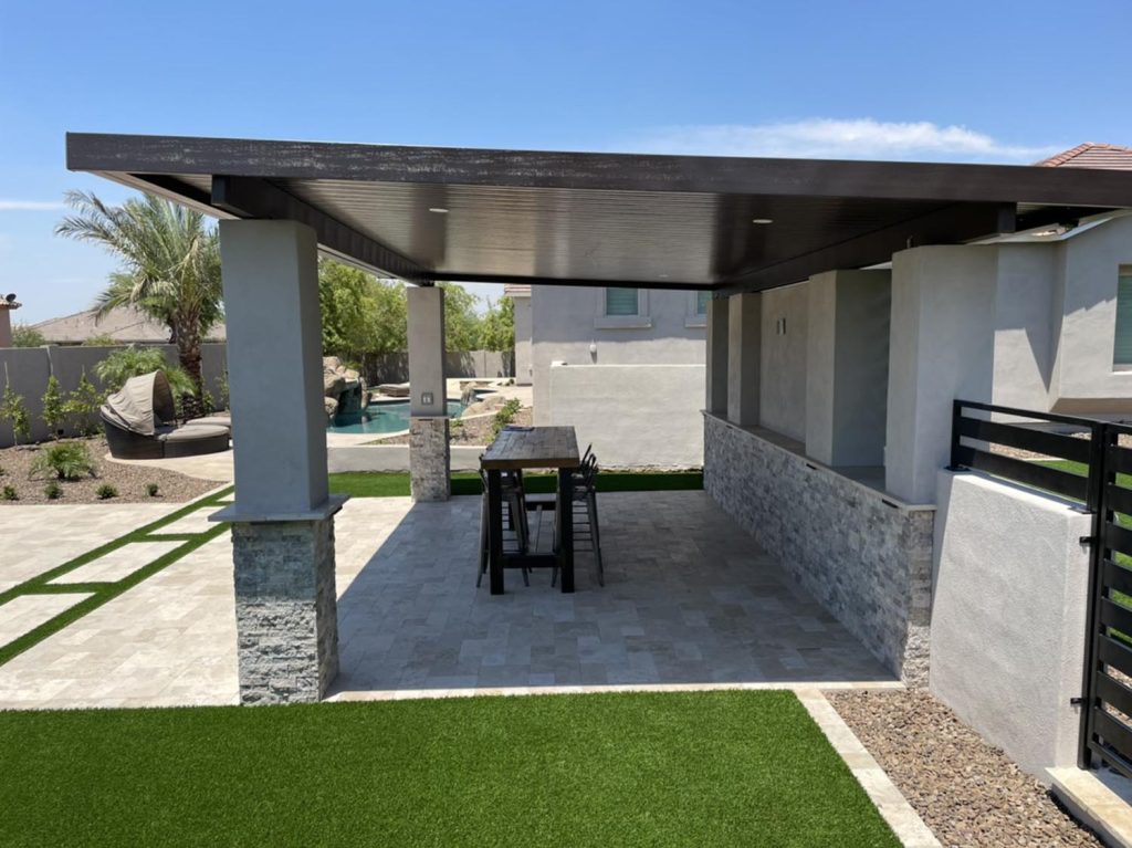 A covered patio area with a dark-colored overhang supported by stone columns, outdoor furniture and a table set on a paver floor, overlooking a yard with synthetic grass and a pool in the distance.