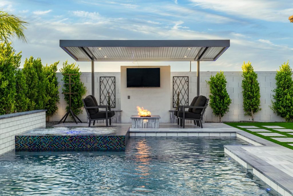 An outdoor living space featuring a roofed area with a flat-screen TV above a fireplace, two armchairs facing a fire pit, and a swimming pool with a mosaic tile feature, bordered by hedges and synthetic lawn at dusk.