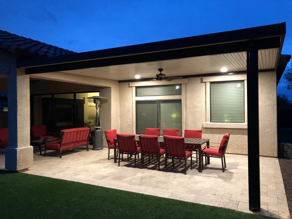 A patio illuminated by overhead recessed lighting with outdoor seating including red cushioned chairs and a bench around a patio table, set against a residential building with large windows and a dark overhang.