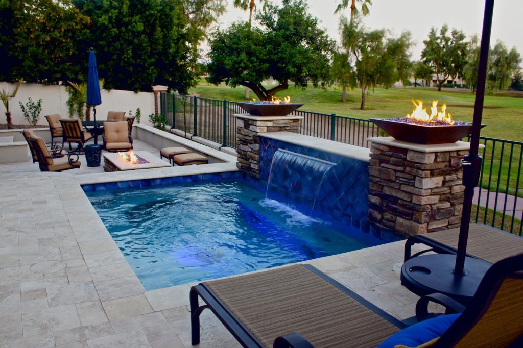 A backyard with a blue-lit swimming pool featuring a cascading waterfall. Adjacent to the pool is a seating area with cushioned chairs and a fire pit table. A large fire bowl stands elevated on a stone pillar. Beyond a metal fence, trees and an open grassy area can be seen.