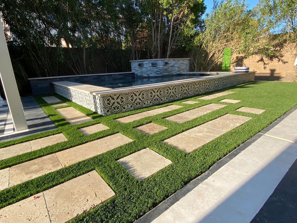 A modern backyard design featuring square stone tiles interspersed with strips of artificial grass. On one side is a raised rectangular water feature with decorative tile sides. Tall trees border the area, and a brown privacy wall is in the background.