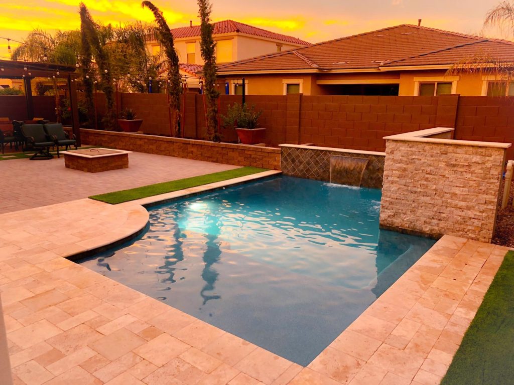 A backyard during sunset with a clear blue pool in the foreground. Beside the pool is a tan-colored paved area and a raised fire pit. There's a seating area under a pergola with hanging lights. Tall palm trees and residential houses are visible in the background under an orange sky.