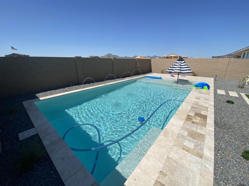 An inviting pool is set against a high gray wall, offering privacy and seclusion. In the distance, mountain ranges stretch across the horizon, while a striped umbrella and floating pool toys hint at leisure and recreation.
