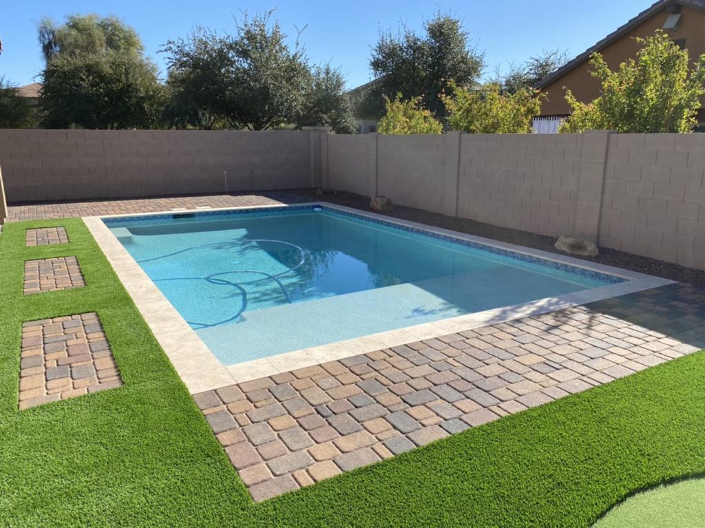 A clear blue pool is highlighted by surrounding multicolored stone tiles and a section of vibrant synthetic grass. A gray brick wall offers privacy, while natural shrubs and trees peek over the barrier, contrasting with the built environment.