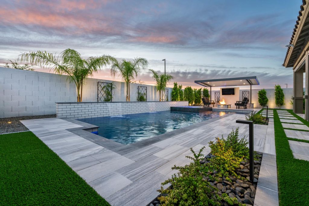 A modern backyard at dusk featuring a swimming pool with blue water, a white brick wall, palm trees, a fire pit area with chairs, and a sunset sky.