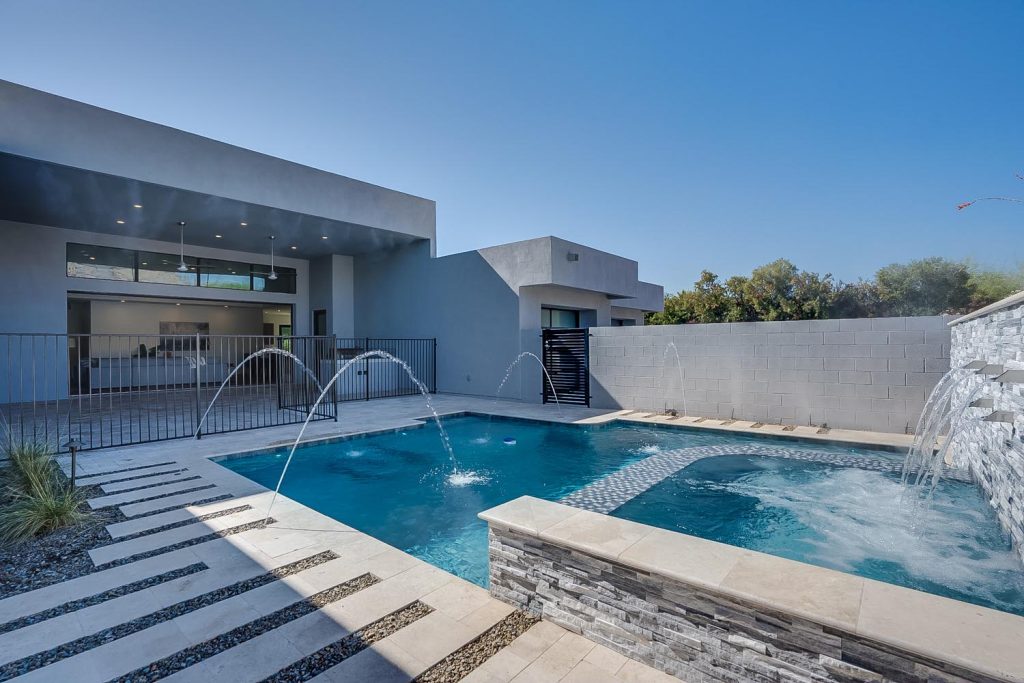 A modern home with a spacious patio featuring a pool with arching water fountains. The house displays large windows and a grayish facade, complemented by a stone pathway and gray walls surrounding the property.