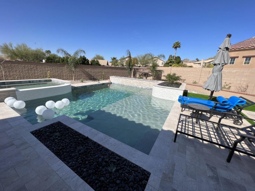 A clear blue swimming pool with floating white balloons, surrounded by a paved patio and a tall brick wall. A hot tub is adjacent to the pool. Nearby, there's a seating area with a blue lounger, black table, and a closed gray umbrella. Palm trees and houses can be seen in the distance.