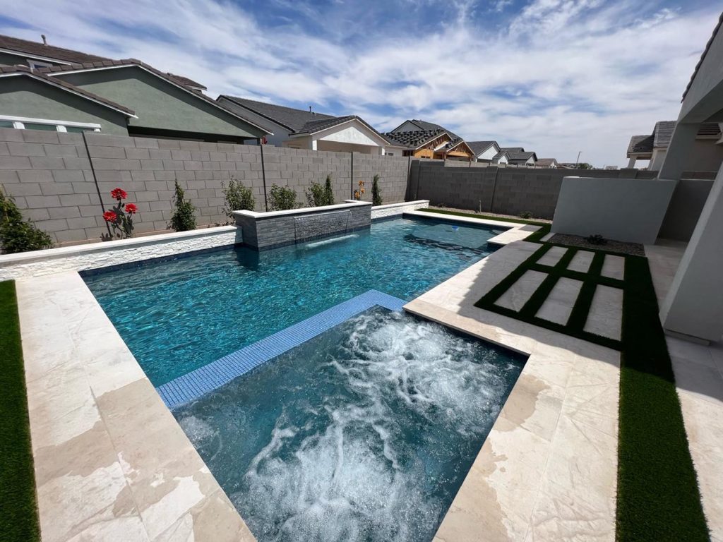 An outdoor residential area featuring a rectangular swimming pool with clear blue water, a cascading waterfall feature on one end, and an adjacent whirlpool with bubbling water. A patterned artificial grass design is laid next to the pool, and the surrounding area is paved with cream-colored tiles. Residential houses with gray roofs are visible over a concrete wall.