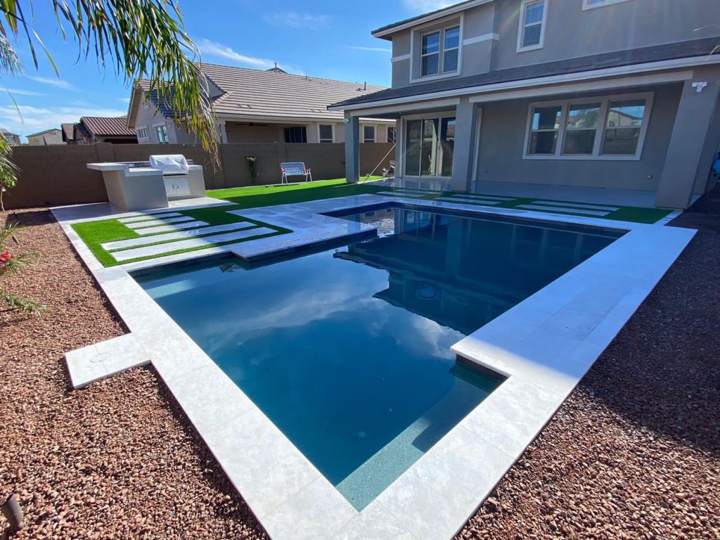 A view of an outdoor residential swimming pool area with a clear blue pool and an extended rectangular design. To one side, there's an outdoor grill or counter, and a patterned artificial grass design. Cream-colored tiles surround the pool. Gravel covers the outer edge of the yard, and a two-story house with large windows stands in the background. A palm leaf hangs overhead in the foreground.