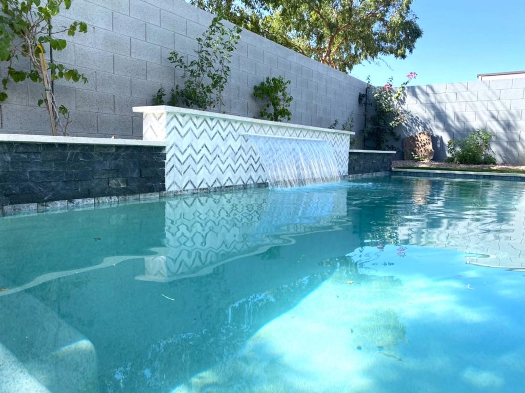 A serene pool setting with clear blue water reflecting sunlight, featuring a white chevron-patterned wall from which water cascades into the pool. Adjacent to the pool, there are trees, flowers, and a gray textured wall providing privacy.