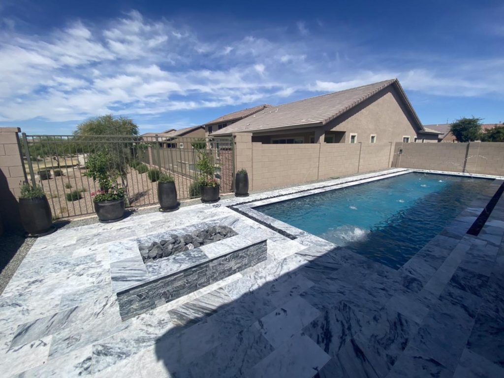A spacious outdoor patio area with a rectangular swimming pool filled with clear blue water. To the side, there's a white and gray marbled seating area with dark pebbles. Beyond the pool fence, a row of houses is visible under a bright blue sky scattered with white clouds. Large planters with plants are aligned against the fence.