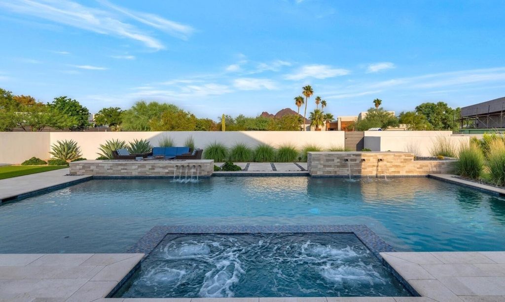 A modern outdoor pool area with a rectangular swimming pool and adjoining hot tub, surrounded by light-colored tiles, stone walls, and native plants, with distant mountains in the horizon.