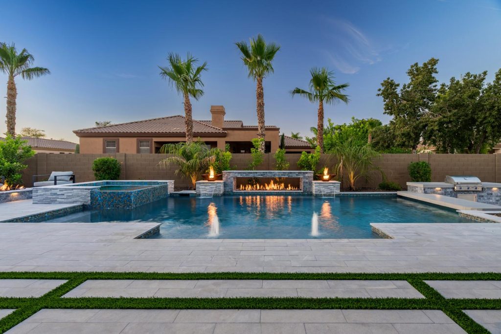 A twilight scene of a backyard featuring a swimming pool reflecting the blue sky, raised mosaic-tiled spa, an elongated fire pit, tall palm trees, and a house with a brown roof, all surrounded by a brick wall.