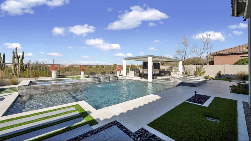 A spacious backyard with a clear blue pool, raised spa with cascading water, patterned synthetic grass sections, tall cacti behind a fence, and an outdoor seating area under a pergola, set against a clear sky.