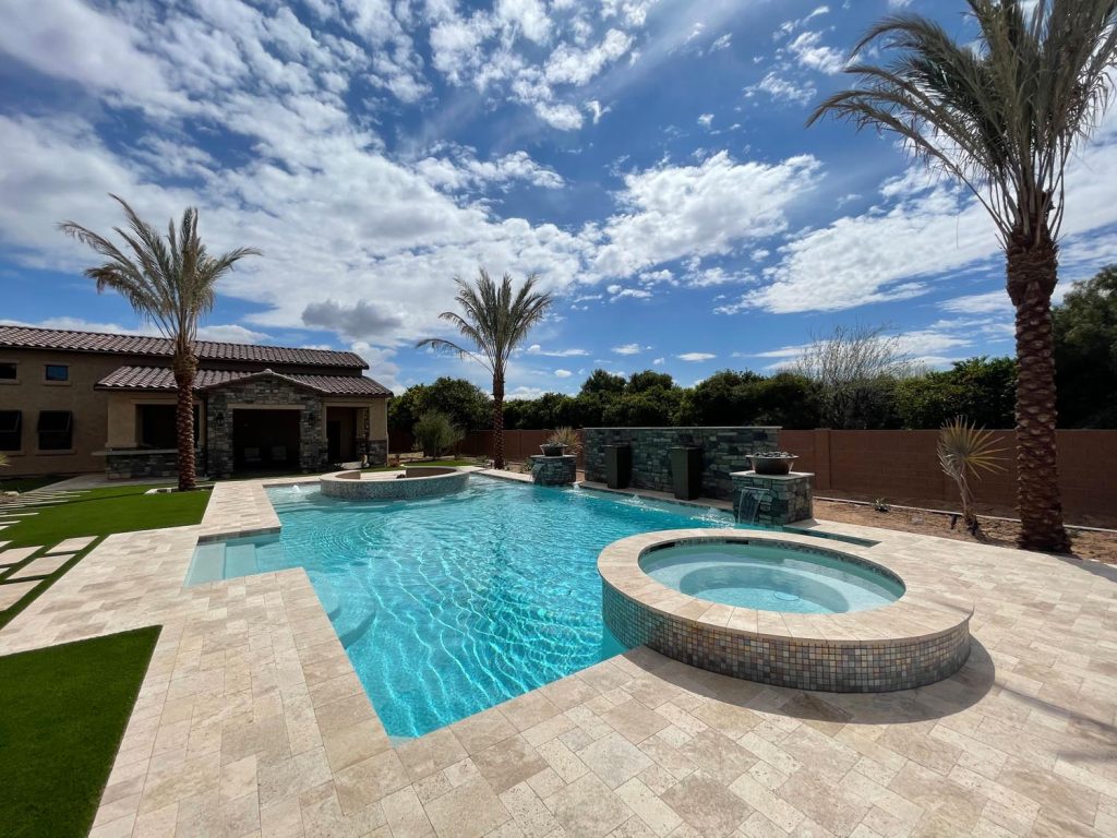 An outdoor residential area featuring a clear blue swimming pool with an attached round hot tub, bordered by pale tiled flooring. A stone-clad house and tall palm trees stand under a vast blue sky dotted with white clouds.