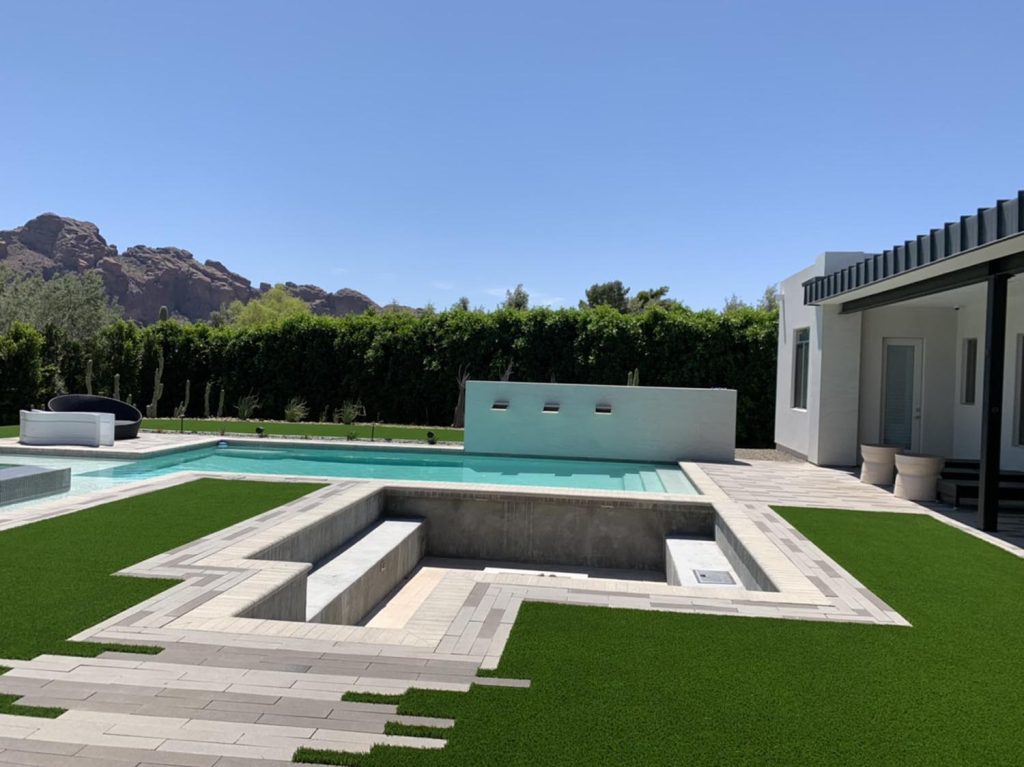 A luxurious outdoor space with a large swimming pool adjacent to a light-colored lounge area. In the distance, rock formations rise against a clear blue sky, while the foreground shows an artificial grass lawn, a raised spa with three waterfall outlets, and a contemporary home facade with large windows.